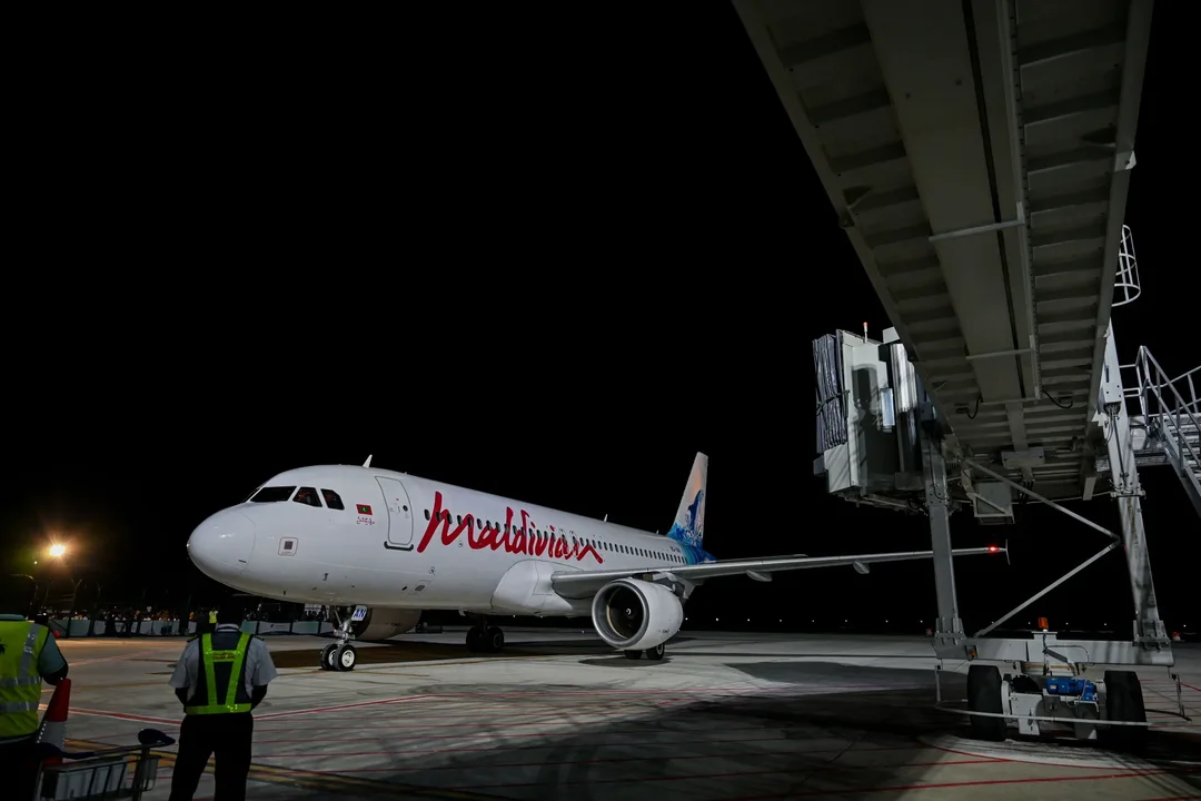 A white Maldivian Airbus A320 with red script lettering stands beside a passenger boarding bridge at night as ground staff in reflective vests watch.