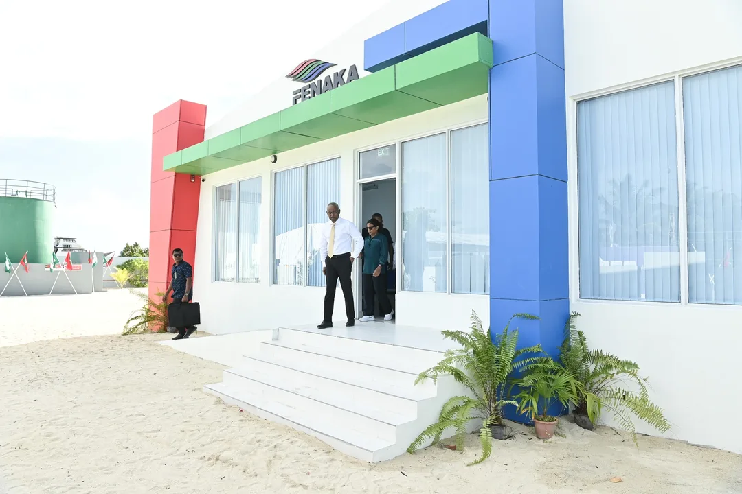 A white single-storey building with red, green and blue panels and a FENAKA sign, with people walking out of its door onto sandy ground.