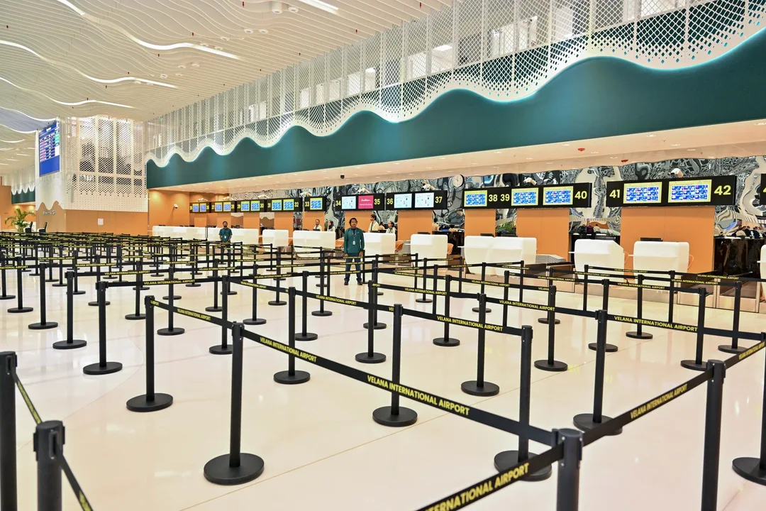 A large airport check-in hall with rows of empty queue barriers in front of numbered check-in counters under a white wave-shaped ceiling.