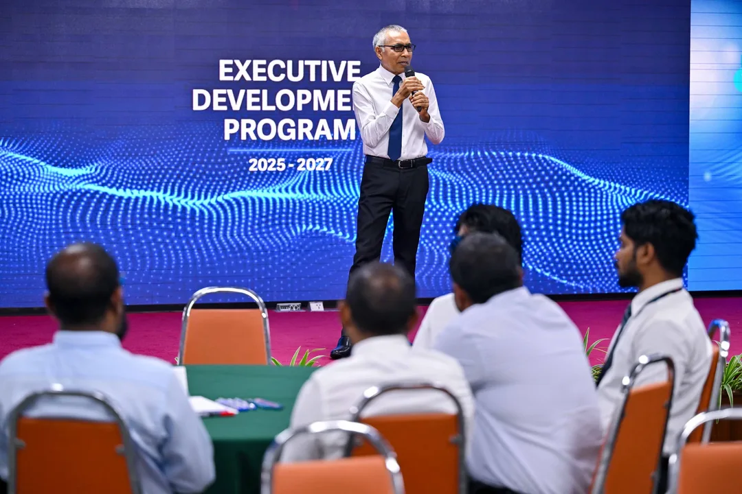 A speaker with a microphone stands before a screen reading Executive Development Program 2025–2027 as officials seated at tables listen.