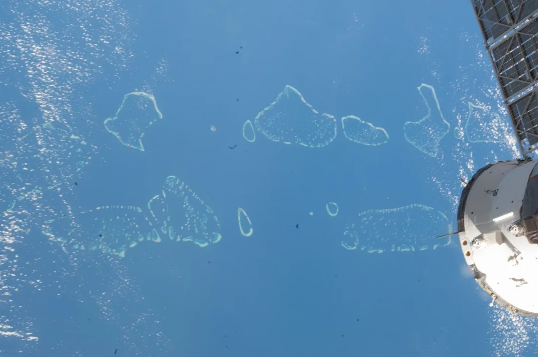 Ring-shaped coral atolls in a blue ocean seen from orbit, with the white hull of a spacecraft and a solar array at the right edge.