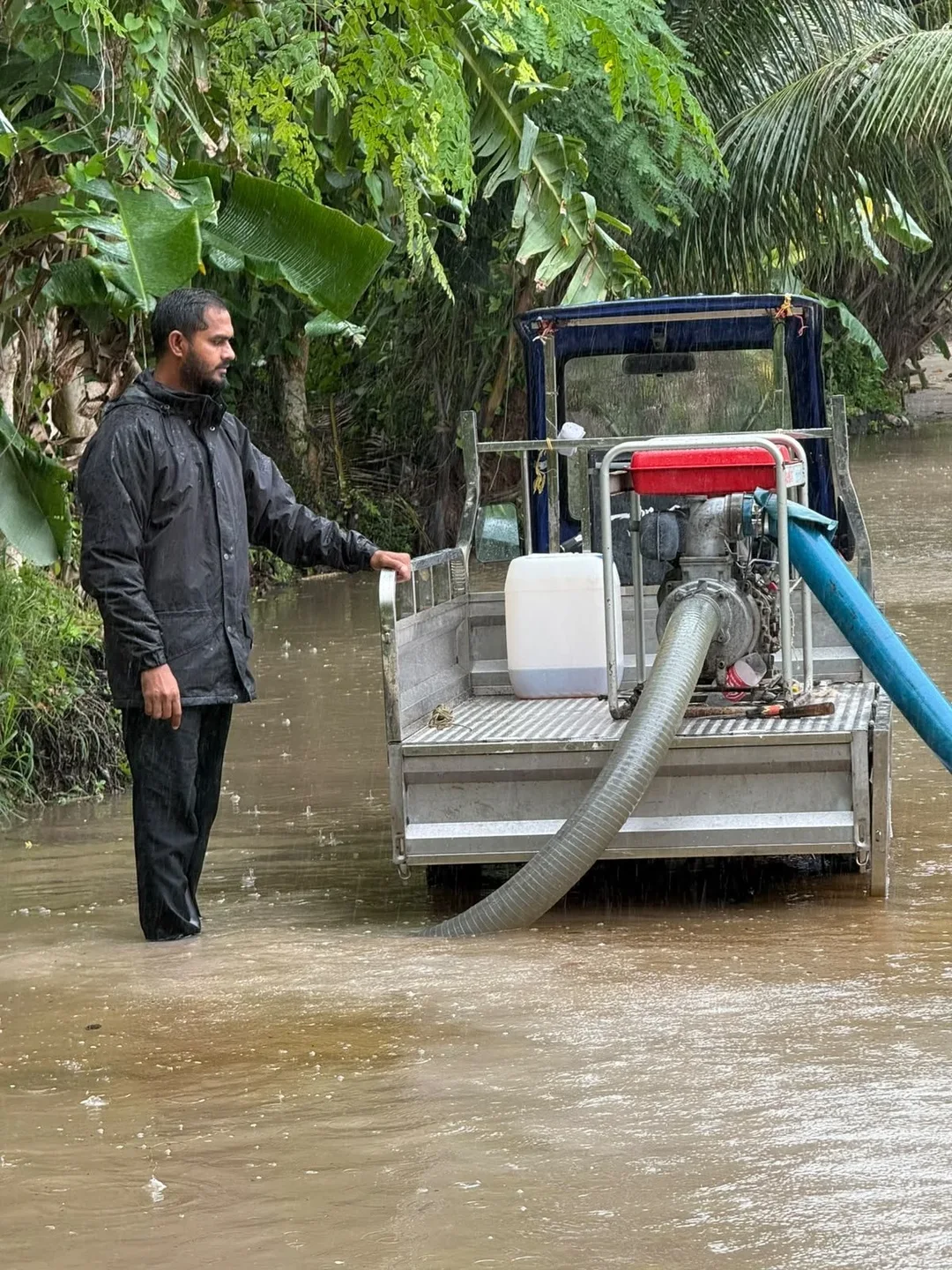 A man in a rain jacket stands in floodwater beside a water pump mounted on a small truck, with a hose running into the water.